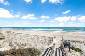 Une plage avec une promenade en bois menant au rivage, des dunes de sable et un ciel bleu dégagé.