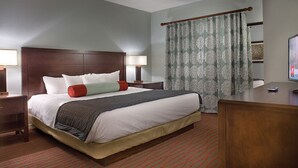 A hotel room with a large bed, two bedside tables with lamps, a desk with a computer, and a window with patterned curtains.