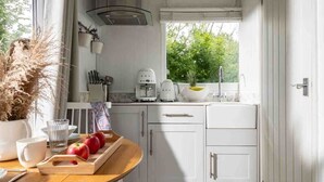 Une cuisine compacte avec des meubles blancs, une table en bois sur laquelle sont disposés des fruits, une machine à café et une fenêtre donnant sur un paysage verdoyant.