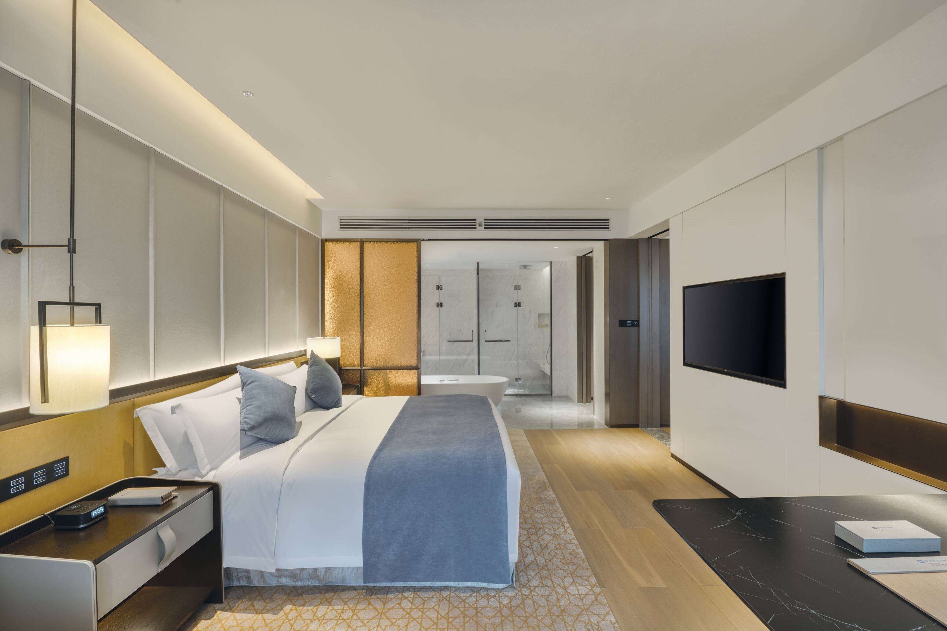 A modern hotel room with a neatly made bed, a flat-screen TV, a bedside table with a lamp, and a large window.