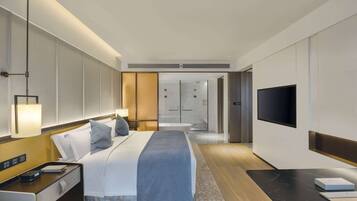A modern hotel room with a neatly made bed, a flat-screen TV, a bedside table with a lamp, and a large window.