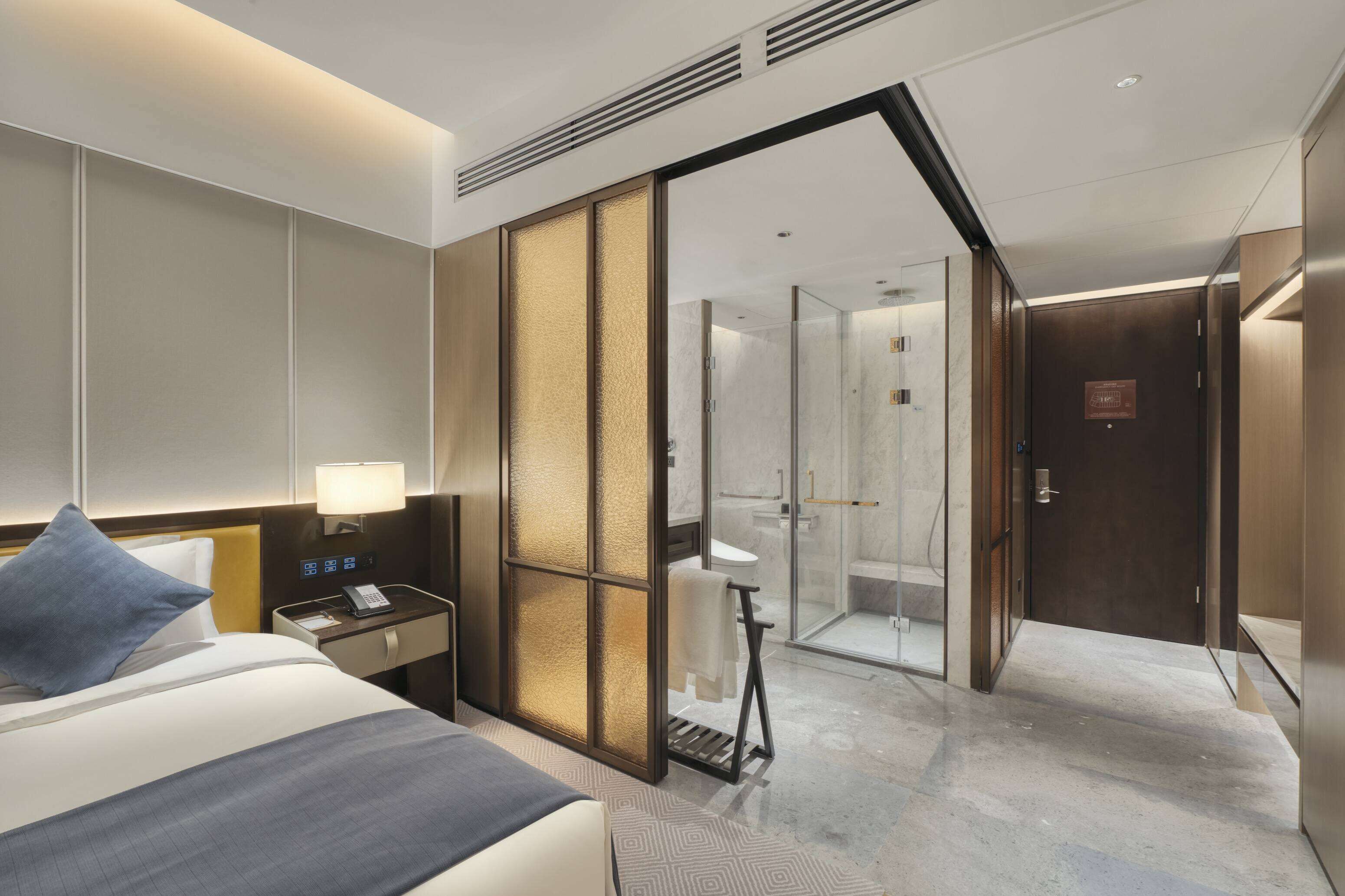 A hotel room with a bed, bedside table, lamp, and a sliding glass door leading to a bathroom with a shower and toilet.