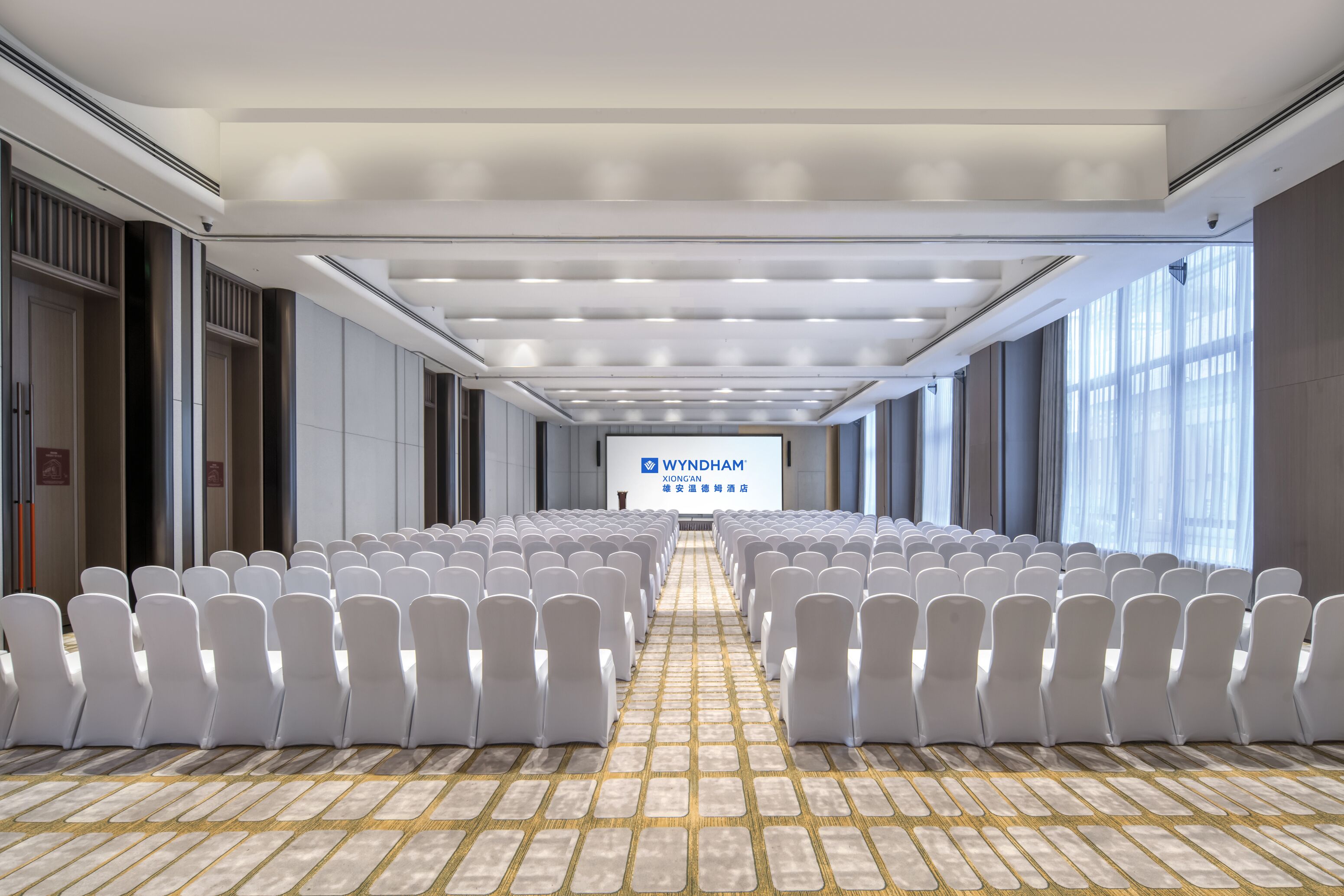 A conference room with rows of white chairs, a tiled floor, and a large screen displaying 'WYNDHAM'.