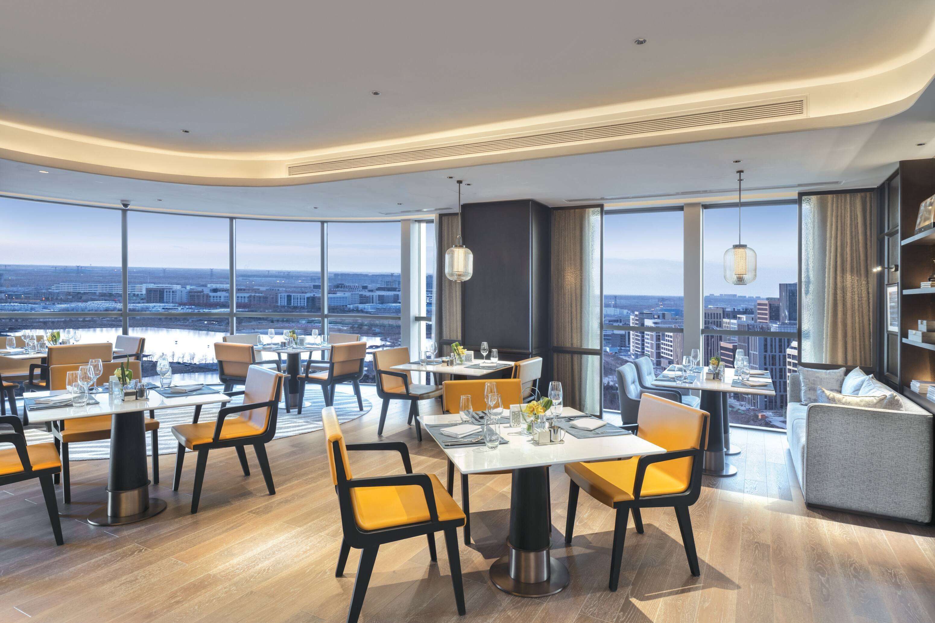 A modern restaurant with high tables and chairs, large windows offering city views, and a cozy seating area with a sofa and side table.