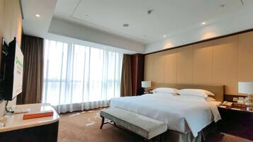 A hotel room with a large bed, bedside lamps, a bench, a desk with a computer, and a window with curtains.