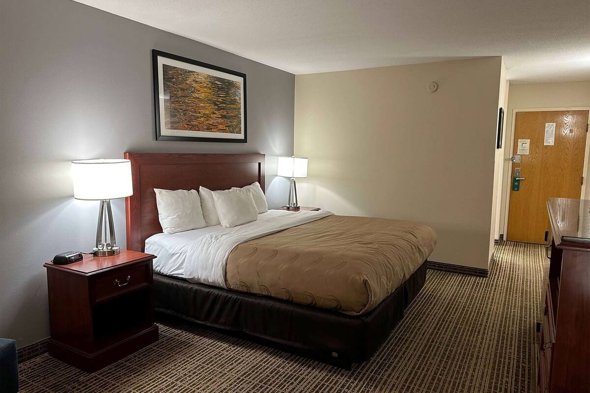 Habitación de hotel con cama, dos mesitas de noche con lámparas, un cuadro en la pared y una puerta a la derecha.