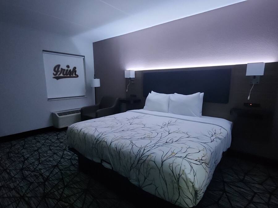 A hotel room with a bed, a chair, a desk, and a framed sign on the wall.