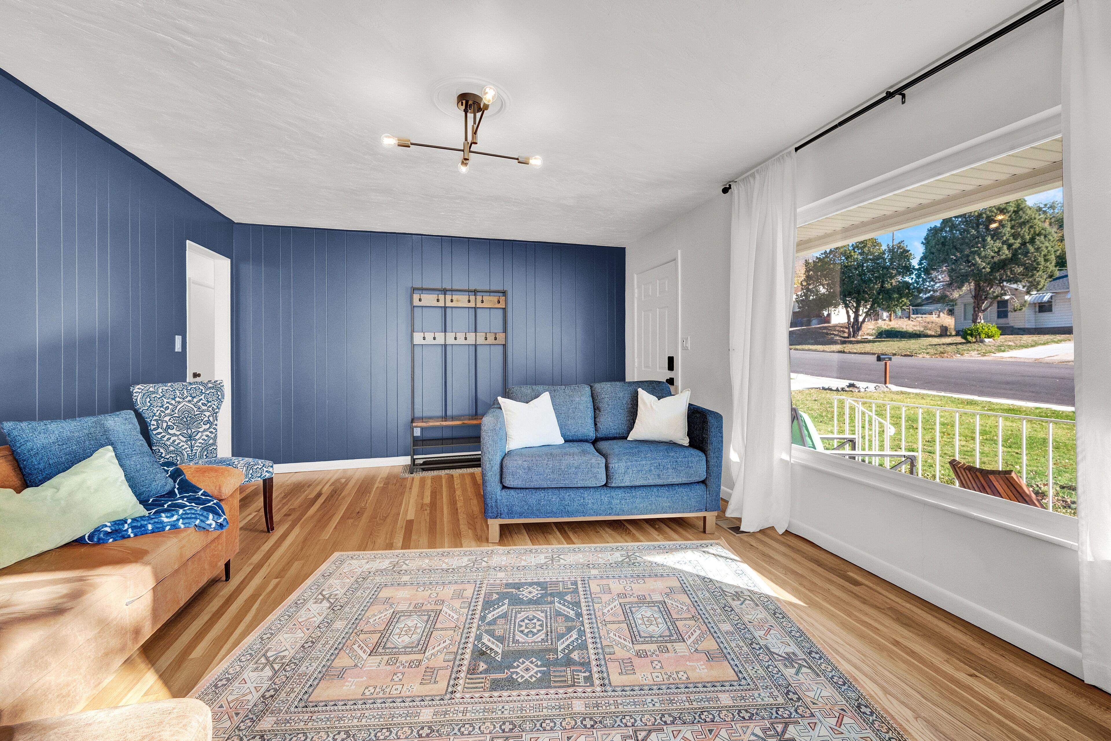 Un salon avec un canapé bleu, un tapis à motifs et une vue sur une rue résidentielle, visible à travers de grandes fenêtres.
