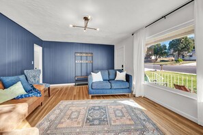 A living room with a blue sofa, a patterned rug, and a view of a residential street through large windows. - Ski Snow Basin | South Ogden  (Ogden)