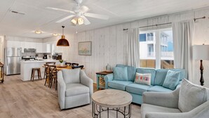 Un vaste espace de vie avec un canapé bleu clair, des fauteuils gris, une table basse en bois et une cuisine en arrière-plan.