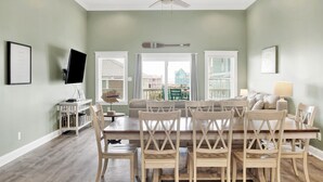 Un comedor con mesa y sillas de madera, un ventilador de techo y vistas a una casa a través de las ventanas.