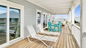 Un porche cubierto con barandillas blancas, una terraza de madera y mobiliario exterior, incluyendo sillones blancos y un juego de mesa y sillas turquesa.