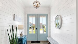 A hallway with a light blue door, a blue and white patterned rug, a potted plant, a blue dresser, and a round mirror on the wall. - Luxury Retreat | Private Pool | Resort Style Amenities (Gulf Shores)