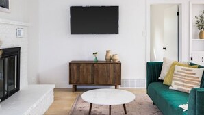 A living room with a fireplace, a round coffee table, a green sofa, and a TV mounted on the wall. - Bright & Modern Retreat - Sleeps 8 - Convenient Location (Wheat Ridge)