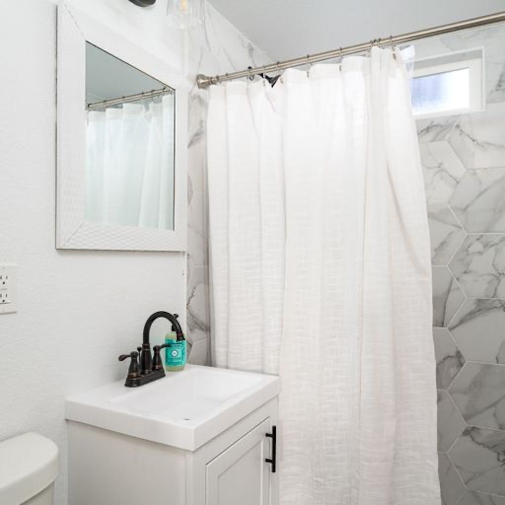 Un baño con lavamanos blanco, un espejo, cortina de ducha y pared con diseño de mármol.
