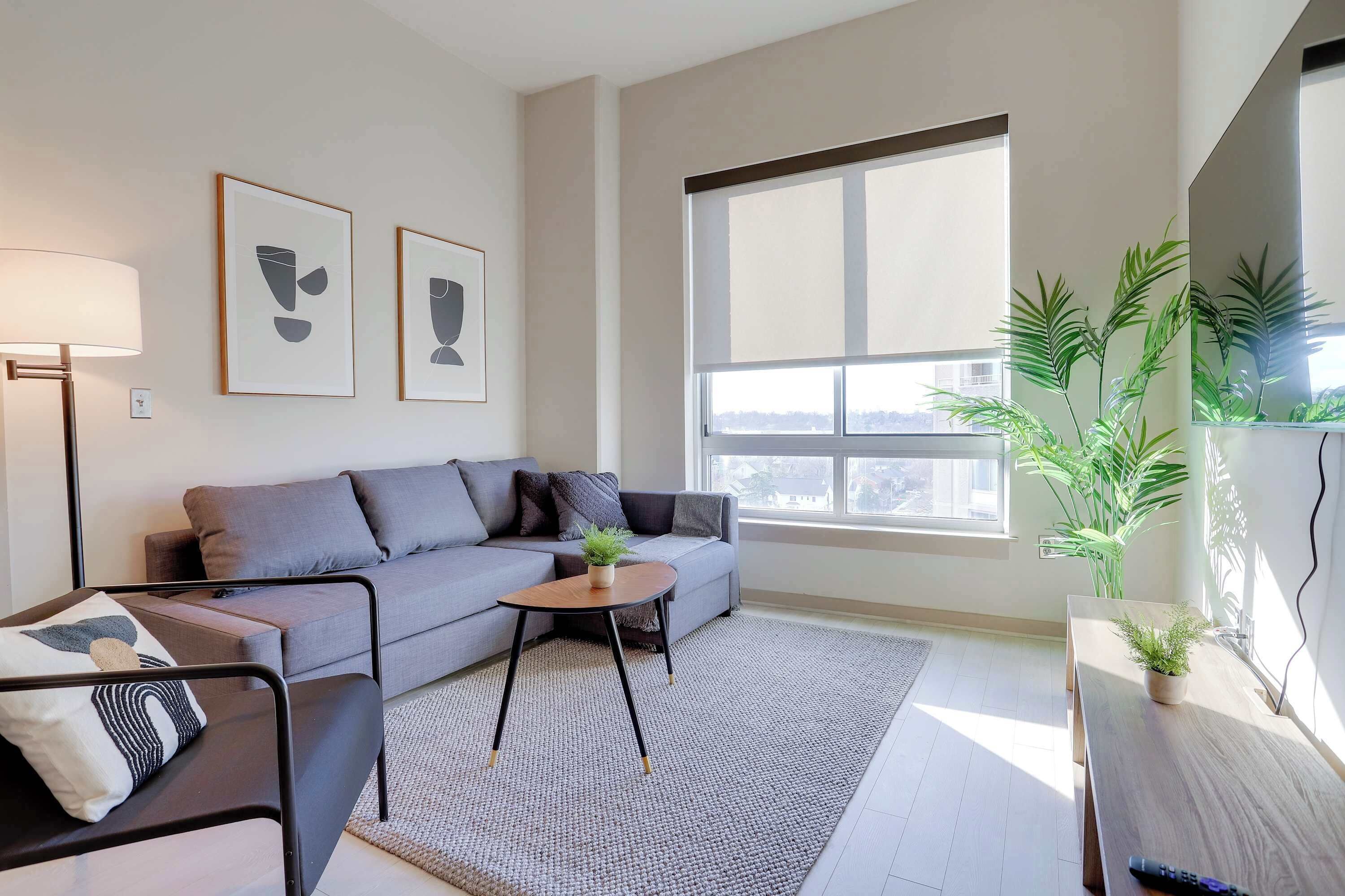 Una sala de estar moderna con un sofá gris, una mesita redonda pequeña, un sillón negro, una lámpara de pie y dos obras de arte abstracto enmarcadas.