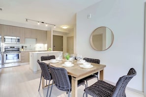 Una cocina moderna con electrodomésticos de acero inoxidable, un comedor con mesa de madera y sillas de mimbre, y un espejo redondo en la pared.