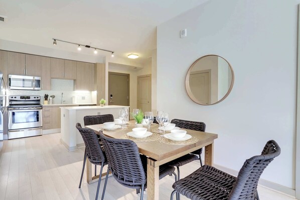 Una cocina moderna con electrodomésticos de acero inoxidable, un comedor con mesa de madera y sillas de mimbre, y un espejo redondo en la pared.