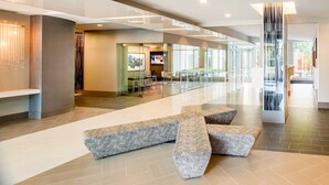 A spacious lobby with a modern design, featuring a central seating area with patterned benches, a glass partition, and a television screen. - Stylish Comfort Meets Spacious Design (Arlington)