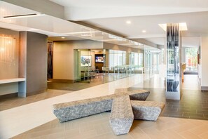 Un lobby espacioso con diseño moderno, que incluye una zona de estar central con bancos estampados, una partición de vidrio y una pantalla de televisión.
