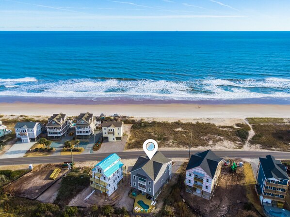 Vista aérea de una zona residencial costera con casas cerca de la playa, una carretera y un icono de ubicación marcado.