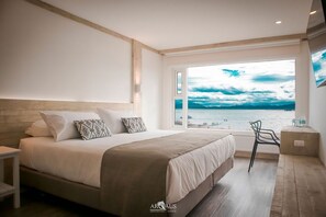 A bedroom with a large bed, a view of the sea, and a chair by the window. - Hotel Costanera (Natales)