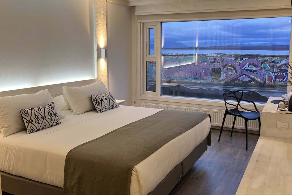 A modern bedroom with a large bed, bedside tables, a chair, and a view of a cityscape through a window.