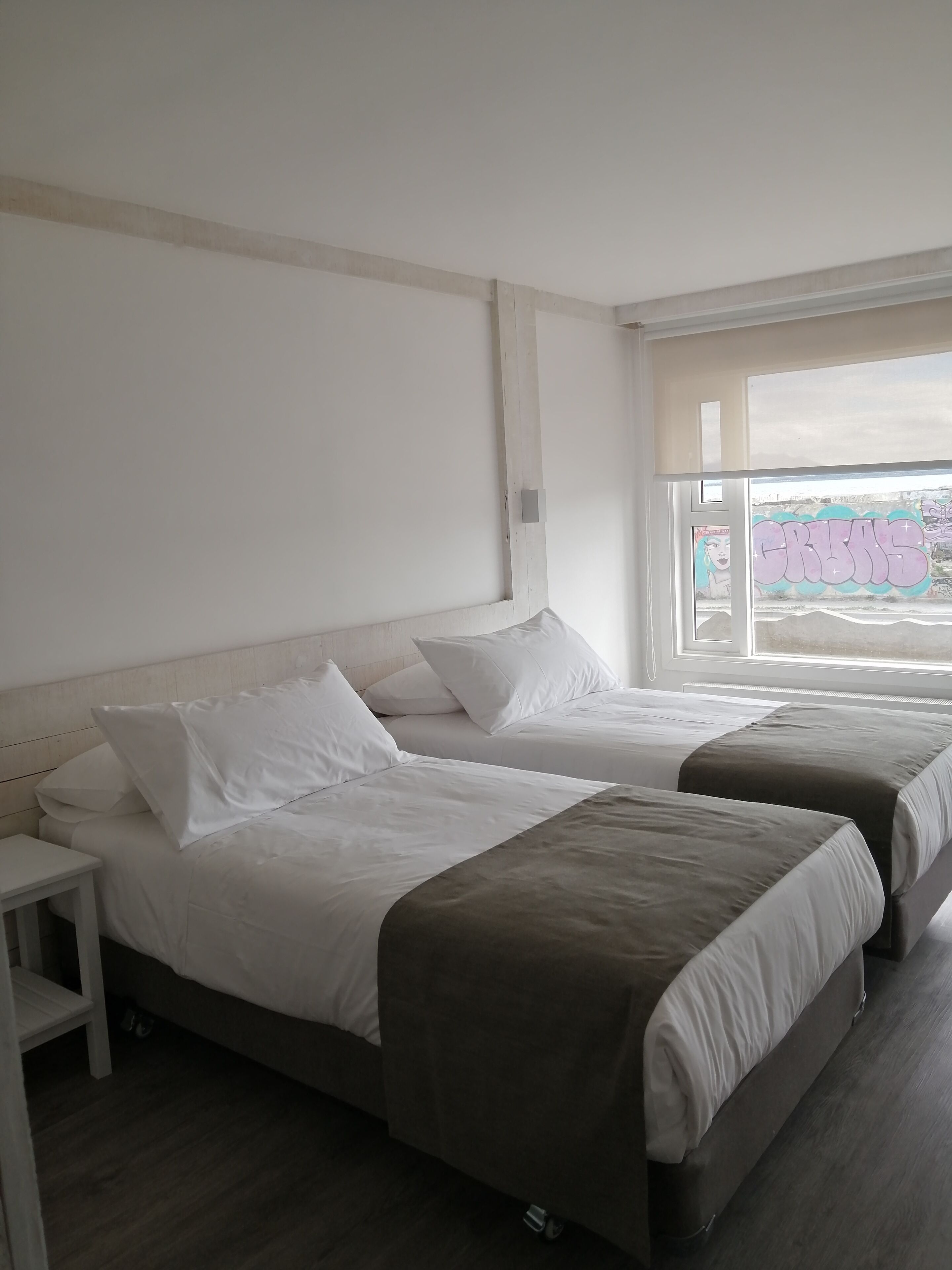 Habitación de hotel con dos camas, una mesita de noche y una ventana con vista a grafitis.