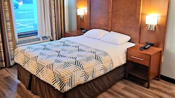 A hotel room with a large bed, wooden headboard, two bedside tables with lamps, a window with curtains, and a view of the outdoors.
