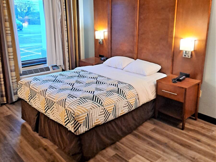 A hotel room with a large bed, wooden headboard, two bedside tables with lamps, a window with curtains, and a view of the outdoors.