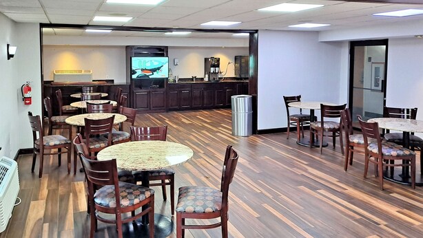 A dining area with round tables and chairs, a kitchen area with a coffee machine, and a fire extinguisher on the wall. - Studio 6 Suites Catonsville, Md – Baltimore West (Catonsville)