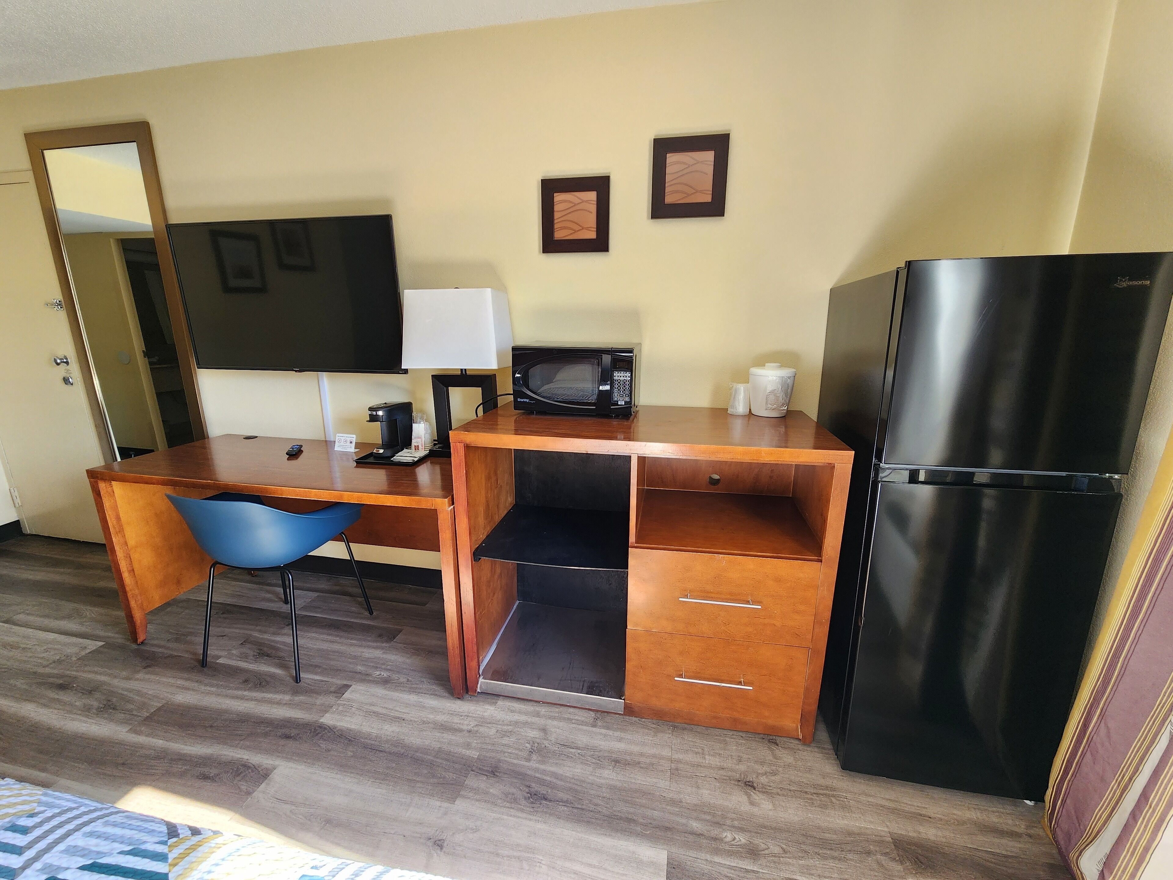 A compact hotel room with a wooden desk, a blue chair, a black refrigerator, a microwave, and a coffee maker.