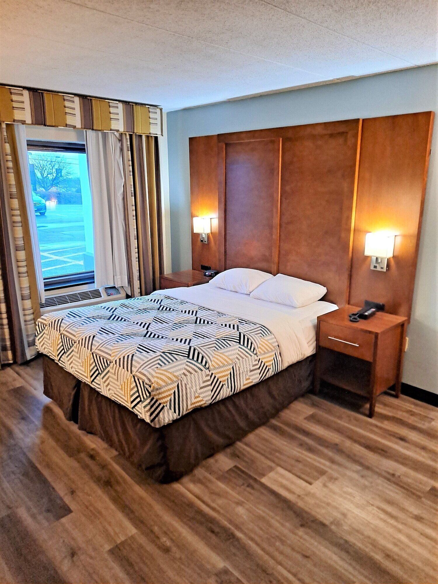 A hotel room with a large bed, wooden headboard, two bedside tables with lamps, and a view of a parking lot through a window.
