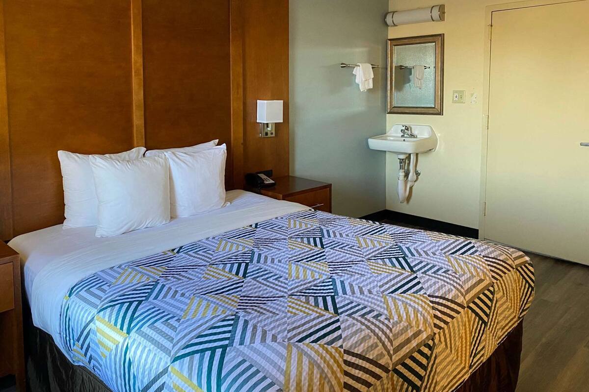 Habitación de hotel con cama, mesita de noche con lámpara, lavamanos empotrado en la pared y una puerta.