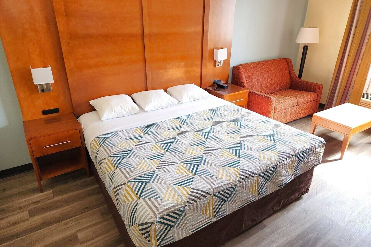 Una habitación de hotel con una cama grande, cabecera de madera, un sillón rojo, una mesita de noche con lámpara y una pequeña mesa.