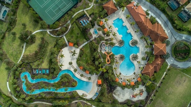 Vista aerea di un resort con una grande piscina centrale, un campo da tennis e una piscina serpentina.