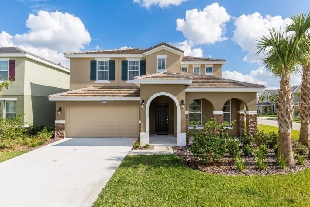 A two-story house with a beige facade, a front garage, and a well-maintained lawn with palm trees.