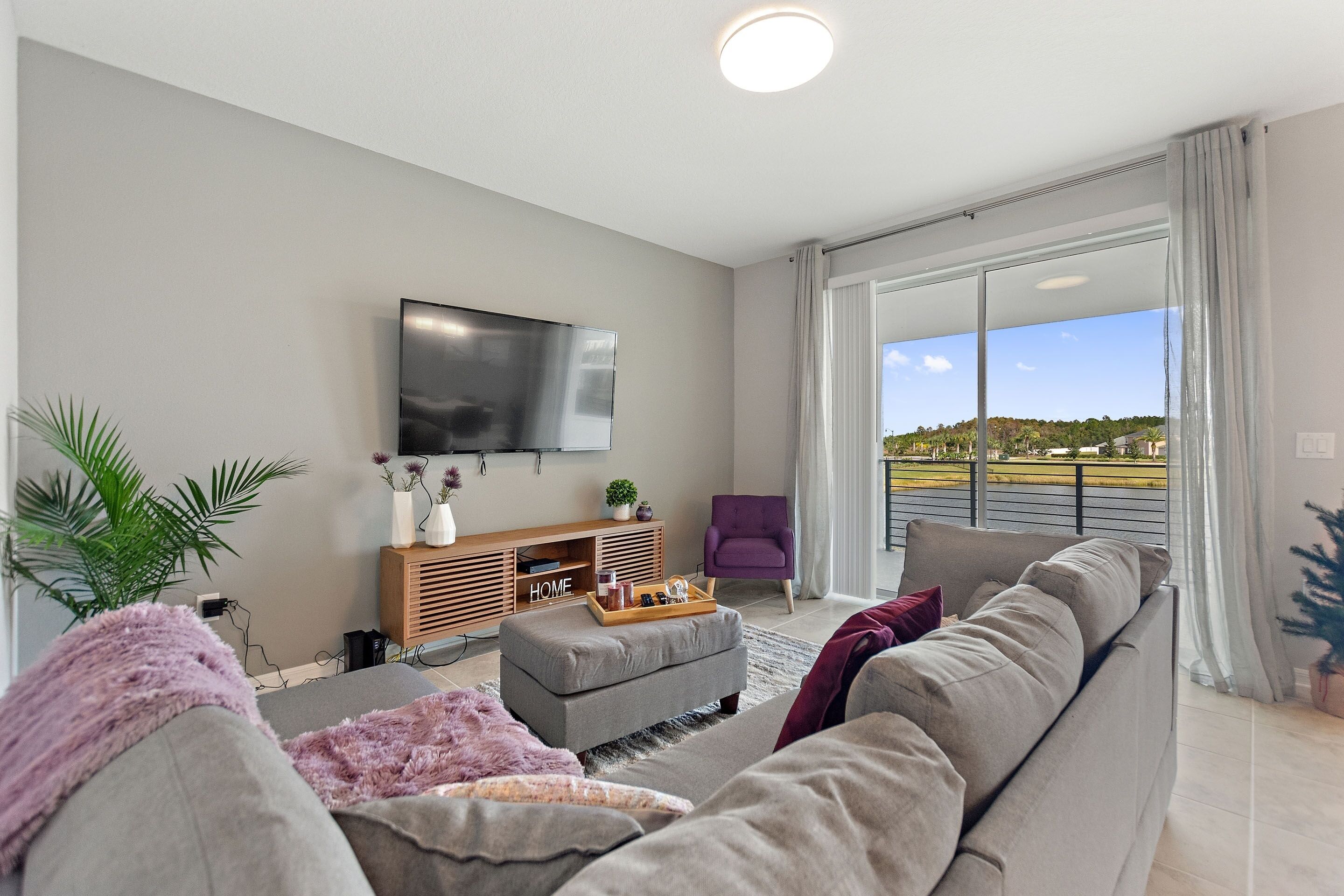 Ein modernes Wohnzimmer mit grauem Sofa, einer Holz-Unterhaltungsecke, einem Flachbildfernseher und einem großen Fenster mit Blick ins Freie.
