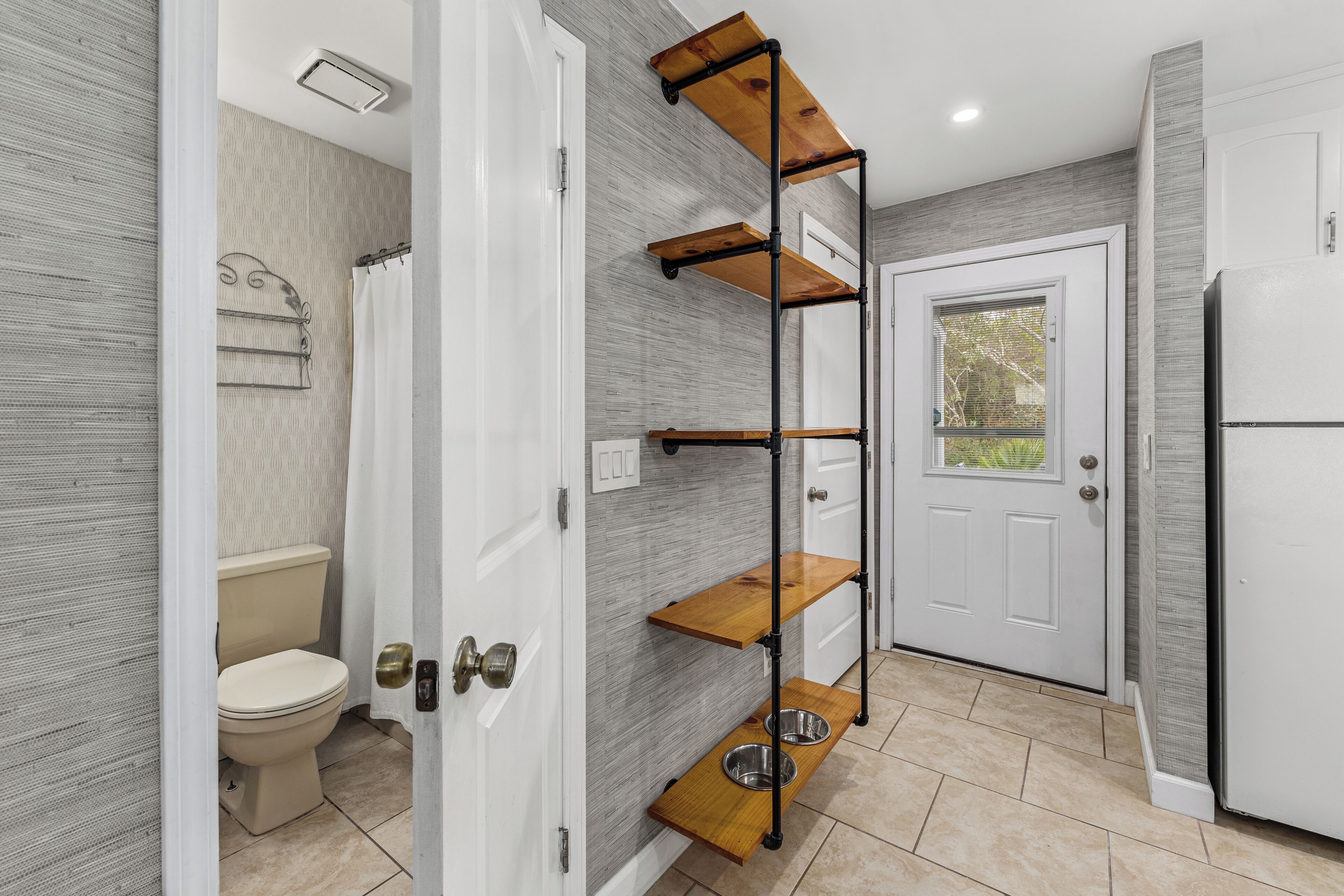A bathroom with a toilet, a shower, and a sink. There is a white door with a window, and a white refrigerator.