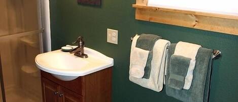 Un baño con un lavamanos blanco, armarios de madera, un perchero con dos toallas, una ventana sobre el lavamanos y una zona de ducha.