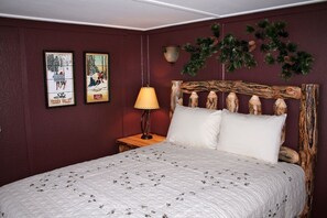 A bedroom with a bed, a nightstand, a lamp, and wall decorations including framed pictures and a pine branch. - Charming Cabin, Next To National Park 3 Bedroom Cabin - #20-NCD0297 (Estes Park)