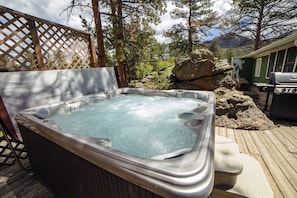 Un jacuzzi en una terraza de madera con vista a árboles y una parrilla al fondo.