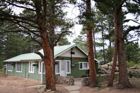 A small, green lodge with a stone entrance, surrounded by tall pine trees. - Charming Cabin, Next To National Park 3 Bedroom Cabin - #20-NCD0297 (Estes Park)
