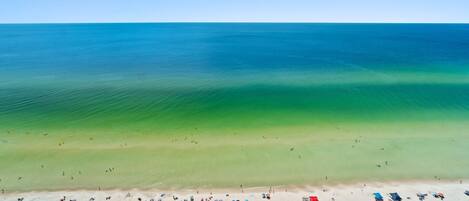 An aerial view of a beach with clear blue water, a sandy shore, and numerous beach umbrellas and chairs.