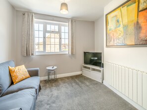 A living room with a sofa, a small table, a TV on a stand, a radiator, and a window with curtains.