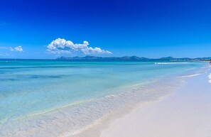 Una playa con agua turquesa cristalina, arena blanca y montañas distantes bajo un cielo azul.