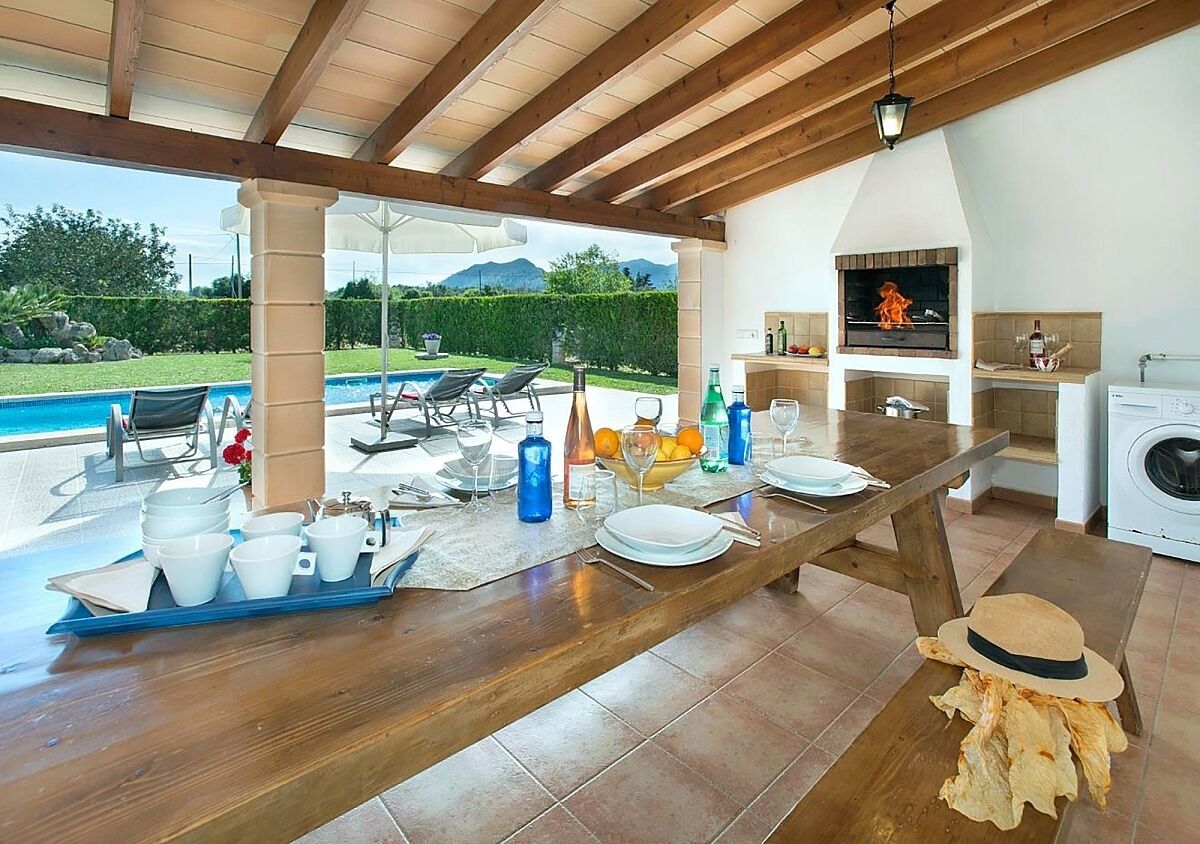 An outdoor dining area with a wooden table set for a meal, a pool, and a kitchen area with a grill.