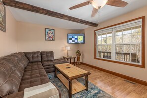 A living room with a sectional sofa, a wooden coffee table, a TV mounted on the wall, and a large window with a view of trees.