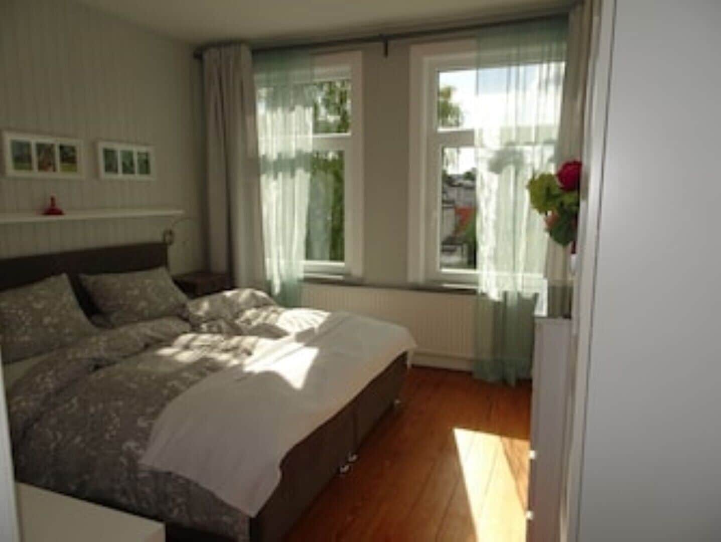 Um quarto com cama, mesa de cabeceira, janela com cortinas e radiador.