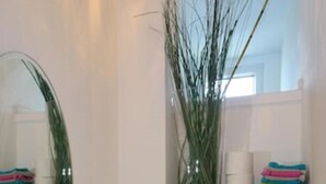 Une salle de bain avec un grand miroir, un lavabo et des toilettes. Une plante est accrochée au mur, et une pile de serviettes colorées y est posée.